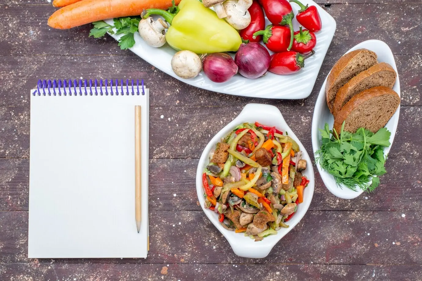 Draufsicht auf frisches Gemüse mit Pilzen in einem Teller, mit Brotlaiben und Grünzeug sowie einem Notizblock auf braunem Hintergrund; Gemüse, Essen, Mahlzeit, Pilz.